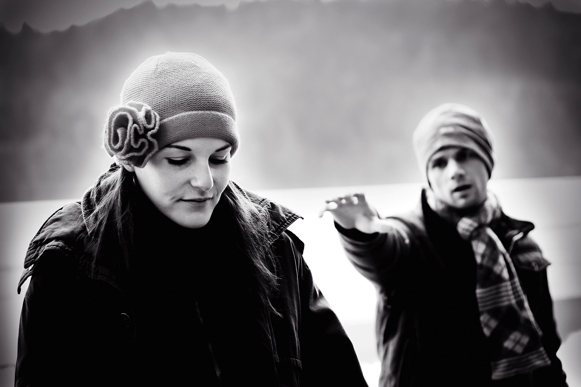 A black-and-white photo of a woman and a man outdoors. The woman, wearing a knit hat with a flower, looks down. The man, in a beanie and scarf, stands behind her with an arm extended. They appear to be in a mountainous area.