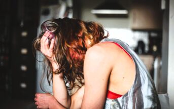 A person with wavy, reddish-brown hair sits with their head in their hand. They are wearing a sleeveless patterned top, and the room is dimly lit, suggesting a kitchen in the background. The person appears distressed or deep in thought.