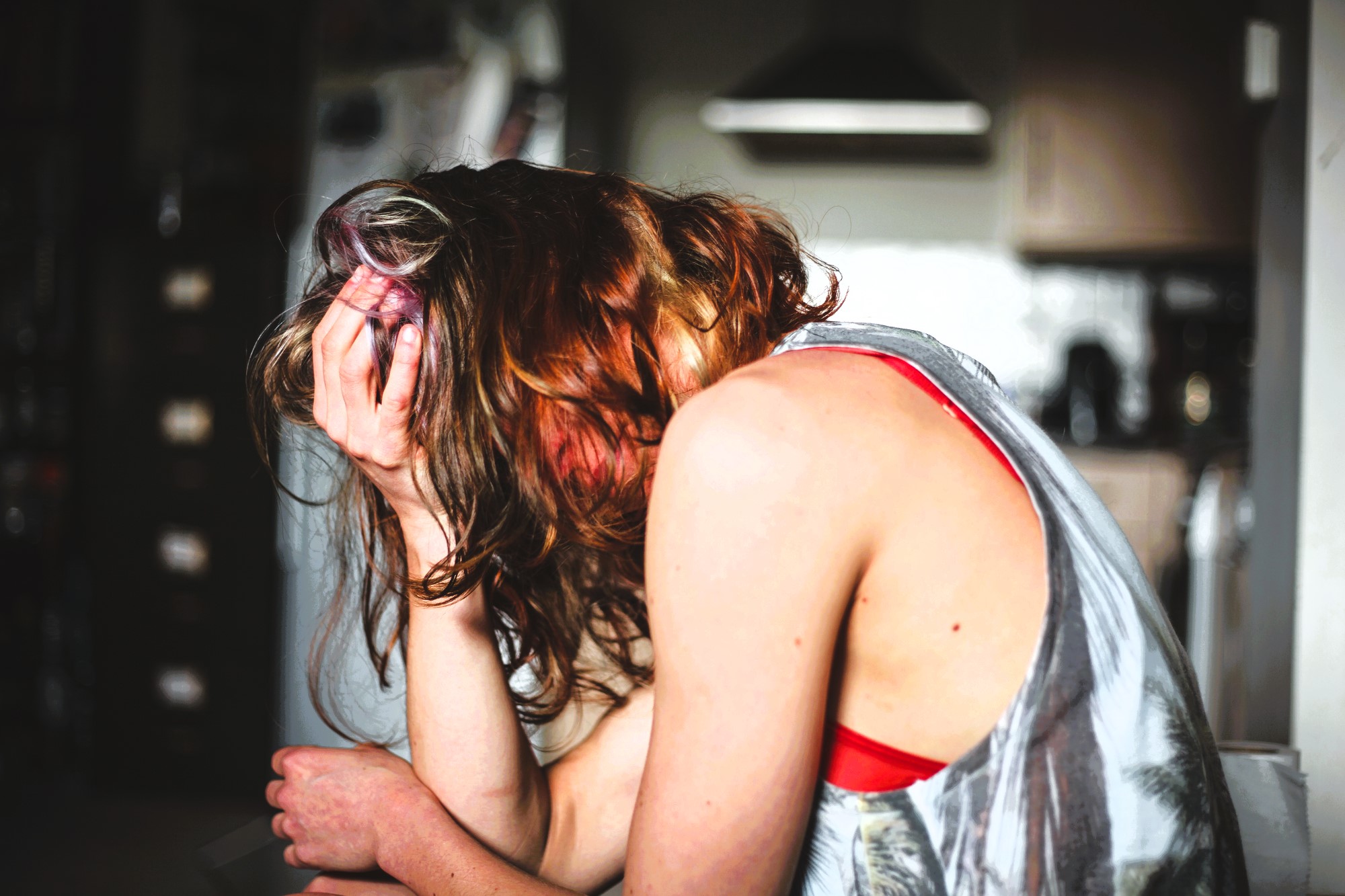 A person with wavy, reddish-brown hair sits with their head in their hand. They are wearing a sleeveless patterned top, and the room is dimly lit, suggesting a kitchen in the background. The person appears distressed or deep in thought.