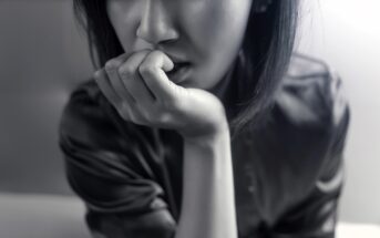 Black and white close-up of a person with shoulder-length hair, sitting and resting their chin on their hand, looking pensive. Their expression is contemplative, with a blurred background.