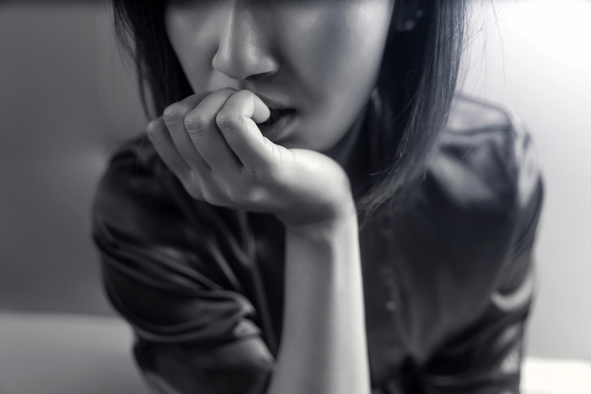 Black and white close-up of a person with shoulder-length hair, sitting and resting their chin on their hand, looking pensive. Their expression is contemplative, with a blurred background.