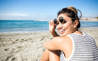 Woman sitting on a sandy beach, smiling and wearing sunglasses and a striped tank top. The ocean is visible in the background, with a clear blue sky overhead.