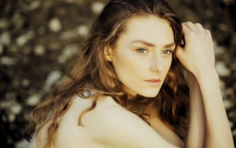 A woman with long, wavy brown hair and light green eyes is sitting outdoors, looking thoughtfully into the distance. She rests her head on her hand, with a soft expression on her face. The background is blurred and earthy.