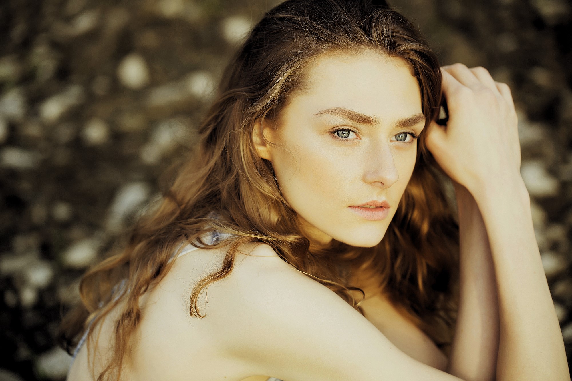 A woman with long, wavy brown hair and light green eyes is sitting outdoors, looking thoughtfully into the distance. She rests her head on her hand, with a soft expression on her face. The background is blurred and earthy.