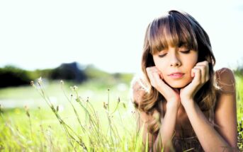 A person with long hair and bangs is lying in a sunny field, resting their head on their hands with eyes closed. The foreground features tall grass and small flowers, while the background is a softly blurred landscape.