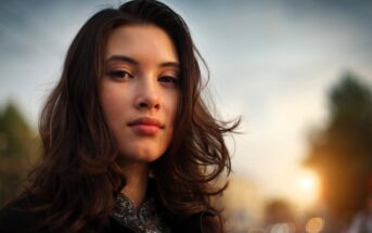 A woman with long, wavy brown hair looks into the camera with a neutral expression. She stands outdoors in soft, warm lighting, with a blurred background of trees and sunset. She's wearing a dark jacket.