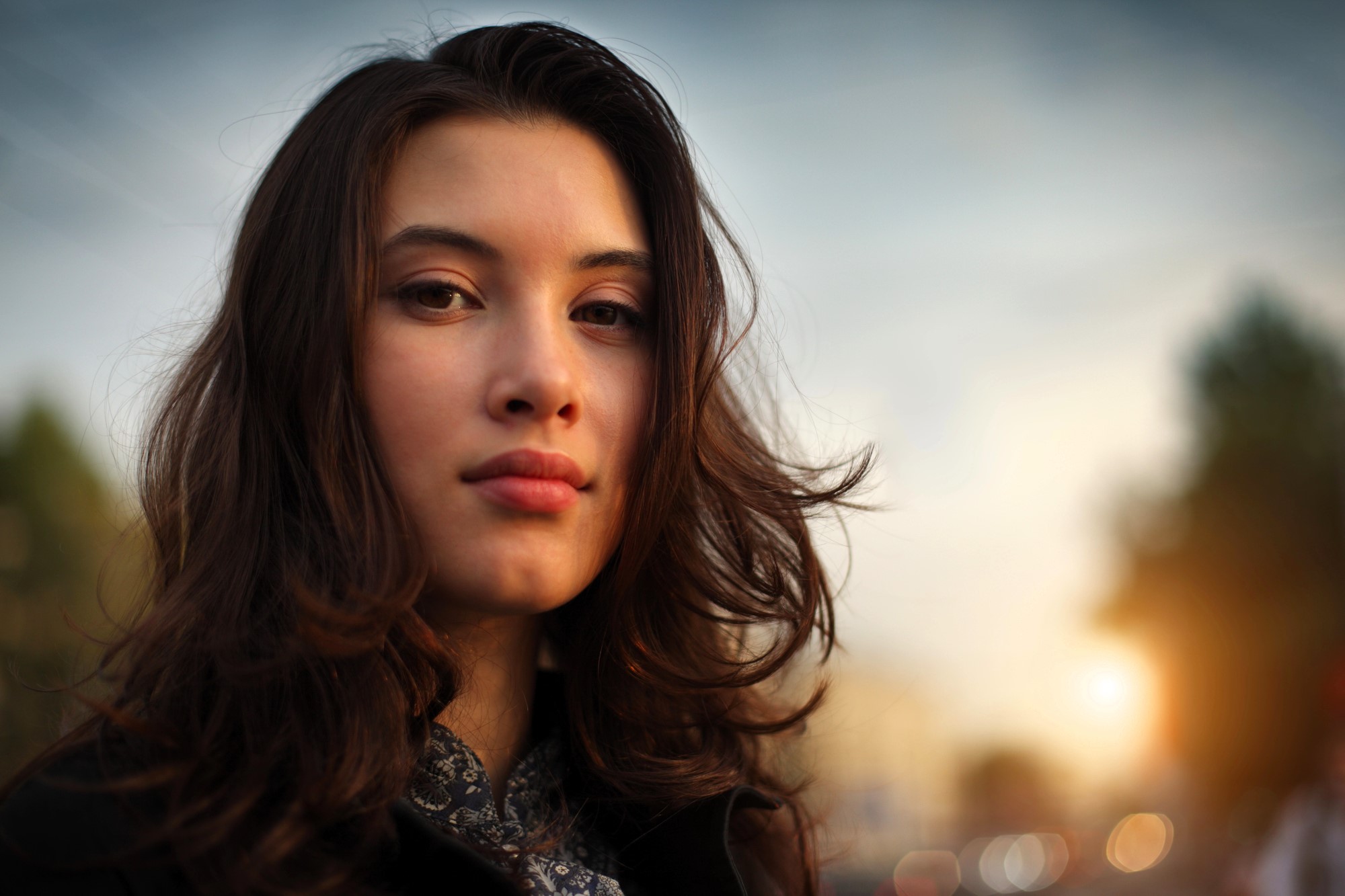 A woman with long, wavy brown hair looks into the camera with a neutral expression. She stands outdoors in soft, warm lighting, with a blurred background of trees and sunset. She's wearing a dark jacket.