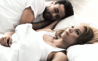 A woman with blonde hair lies awake in bed, looking thoughtful, while a man with dark hair sleeps beside her. They are both on white pillows, covered with a white blanket in a softly lit room.