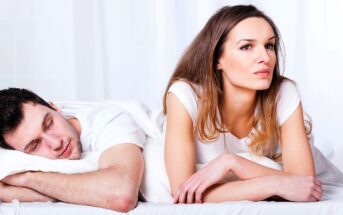 A woman is lying on a bed, propped up on her elbows, looking thoughtful. Next to her, a man is sleeping peacefully with his head resting on his arms. Both are wearing white shirts and the bedding is white.