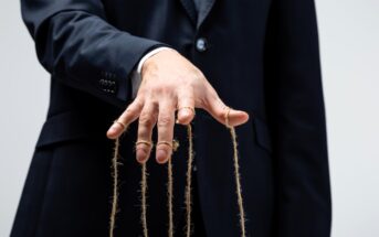 A person in a dark suit stands against a plain background, with their hand extended forward. Strings are attached to their fingers, resembling a puppeteer's control. The image suggests the concept of manipulation or control.