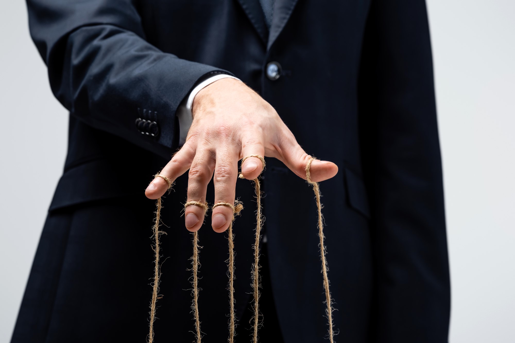 A person in a dark suit stands against a plain background, with their hand extended forward. Strings are attached to their fingers, resembling a puppeteer's control. The image suggests the concept of manipulation or control.