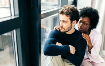 A man sits by a window with a pensive expression, hugging his knees. A woman sits beside him, comforting him with her hand on his shoulder. Both look towards the window, suggesting a moment of support and contemplation.