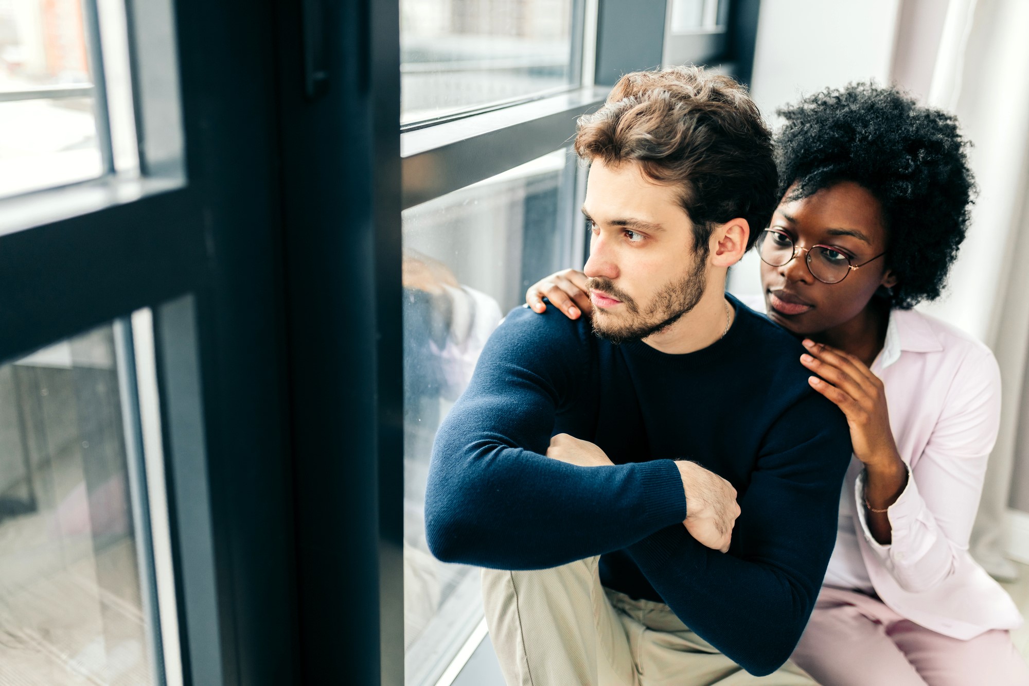 A man sits by a window with a pensive expression, hugging his knees. A woman sits beside him, comforting him with her hand on his shoulder. Both look towards the window, suggesting a moment of support and contemplation.