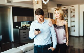 A man and woman are standing in a modern living room. The man, in a light blue shirt and dark pants, looks at his phone. The woman, in a pink camisole and black skirt, rests her hand on his shoulder, looking into the distance.