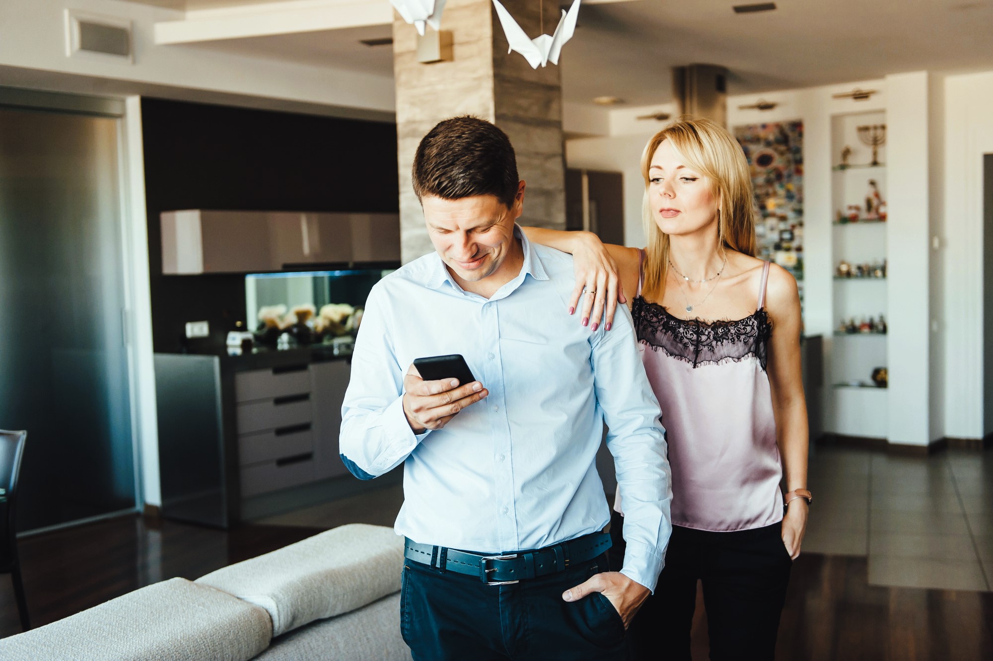 A man and woman are standing in a modern living room. The man, in a light blue shirt and dark pants, looks at his phone. The woman, in a pink camisole and black skirt, rests her hand on his shoulder, looking into the distance.