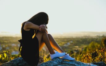 A woman in a black dress sits on a rock, resting her head on her arms, with a serene landscape in the background. The sun is setting, casting a golden hue over the scene. She is wearing white sneakers.