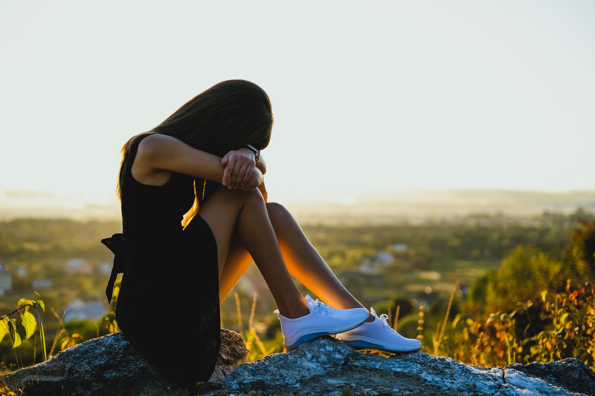 A woman in a black dress sits on a rock, resting her head on her arms, with a serene landscape in the background. The sun is setting, casting a golden hue over the scene. She is wearing white sneakers.