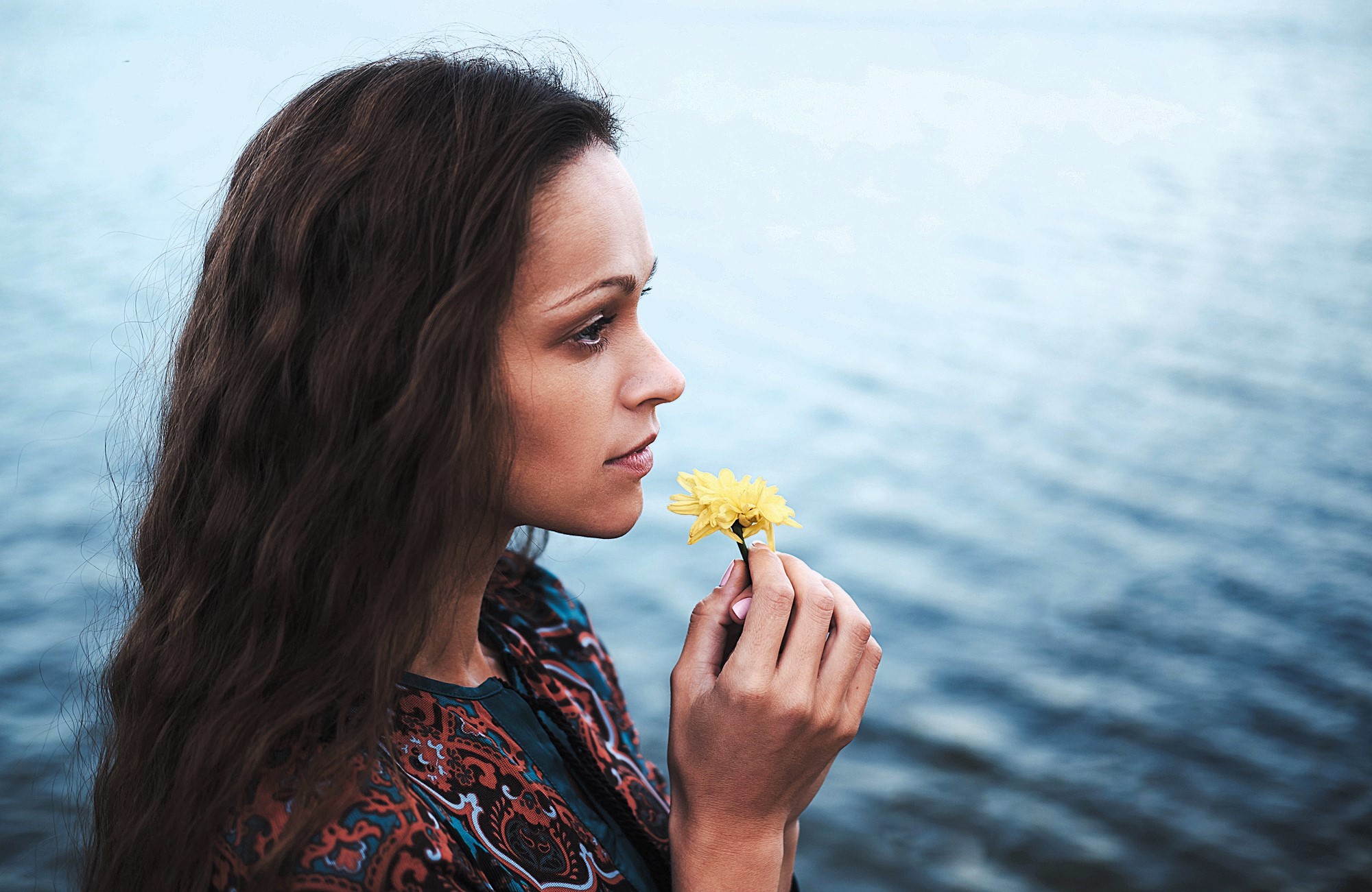 A woman with long brown hair is holding a yellow flower close to her face, gazing thoughtfully at the calm water in the background. She wears a patterned top, and the scene conveys a sense of tranquility.