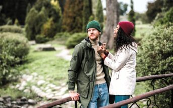 A man and woman in warm clothing stand on a wooden bridge in a park. The woman is smiling at the man, holding his hands. He leans against the railing, looking away. Both wear beanies; hers is red, his is green. Lush greenery surrounds them.