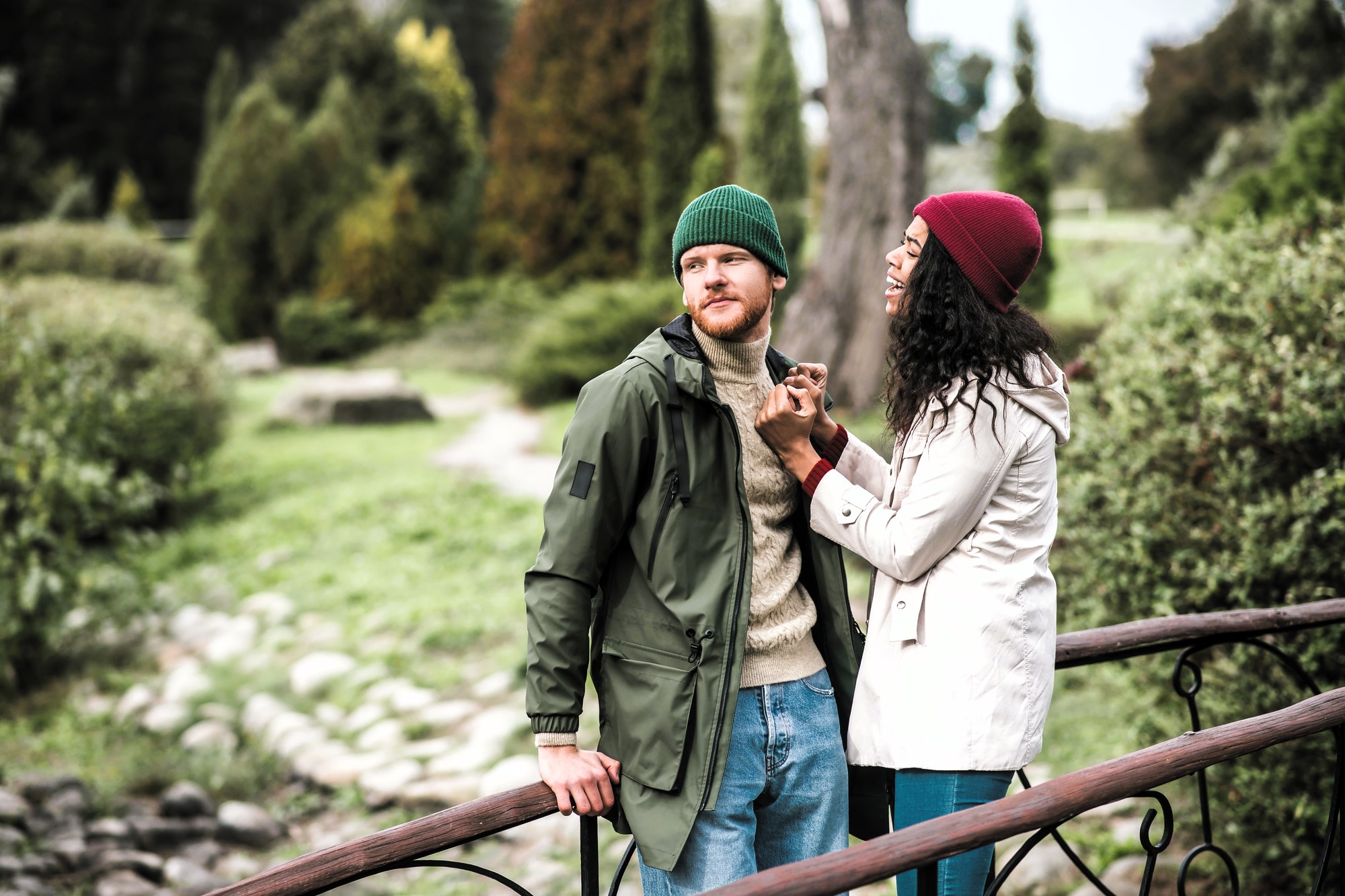 A man and woman in warm clothing stand on a wooden bridge in a park. The woman is smiling at the man, holding his hands. He leans against the railing, looking away. Both wear beanies; hers is red, his is green. Lush greenery surrounds them.