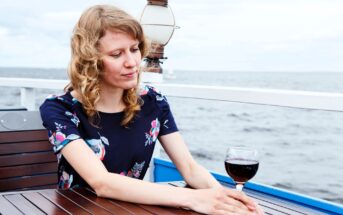 A woman with curly hair sits at a wooden table on a deck overlooking the ocean. She is wearing a floral dress and gazes at a glass of red wine in front of her. The background features a cloudy sky and a nautical lantern.