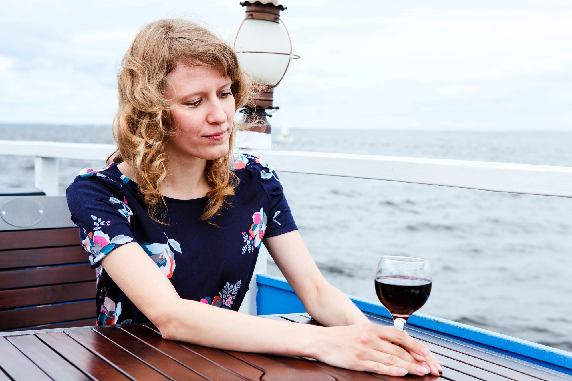 A woman with curly hair sits at a wooden table on a deck overlooking the ocean. She is wearing a floral dress and gazes at a glass of red wine in front of her. The background features a cloudy sky and a nautical lantern.