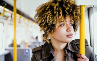 A person with curly hair and a thoughtful expression stands on public transportation, holding a yellow pole. They wear a brown jacket. The background shows an empty bus or train interior.