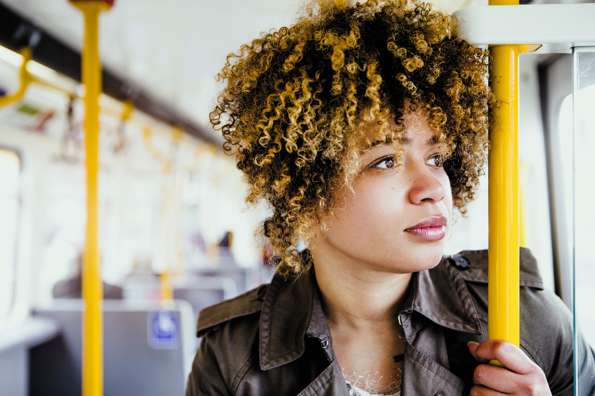 A person with curly hair and a thoughtful expression stands on public transportation, holding a yellow pole. They wear a brown jacket. The background shows an empty bus or train interior.