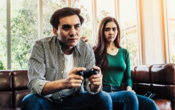 A man and a woman sit on a leather sofa. The man, focused, is holding a video game controller. The woman next to him looks on with a neutral expression. They are in a bright room with large windows and trees visible outside.