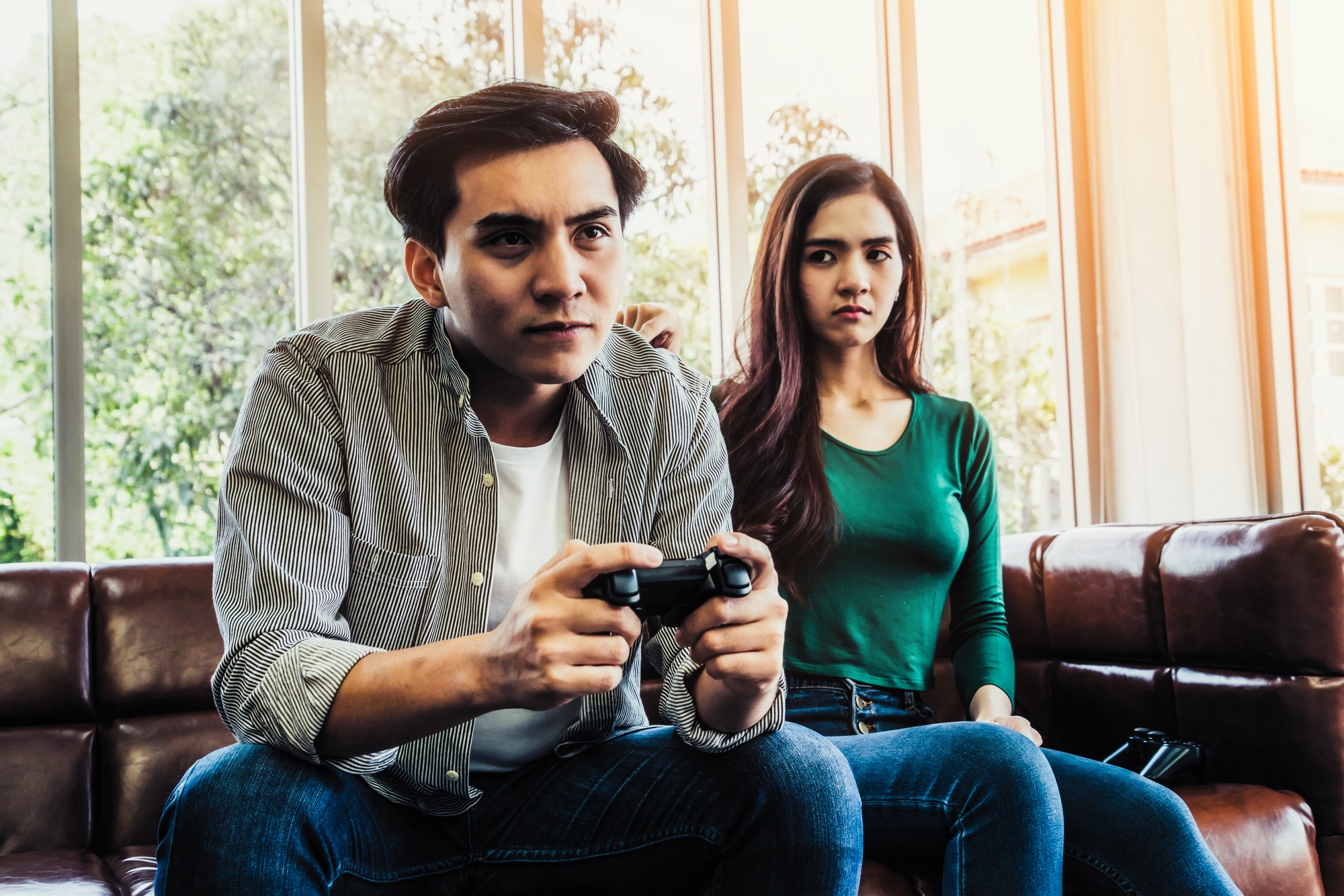 A man and a woman sit on a leather sofa. The man, focused, is holding a video game controller. The woman next to him looks on with a neutral expression. They are in a bright room with large windows and trees visible outside.