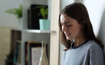 A woman in a striped shirt stands against a wall, looking concerned. The background features a bookshelf with plants and decorative items.