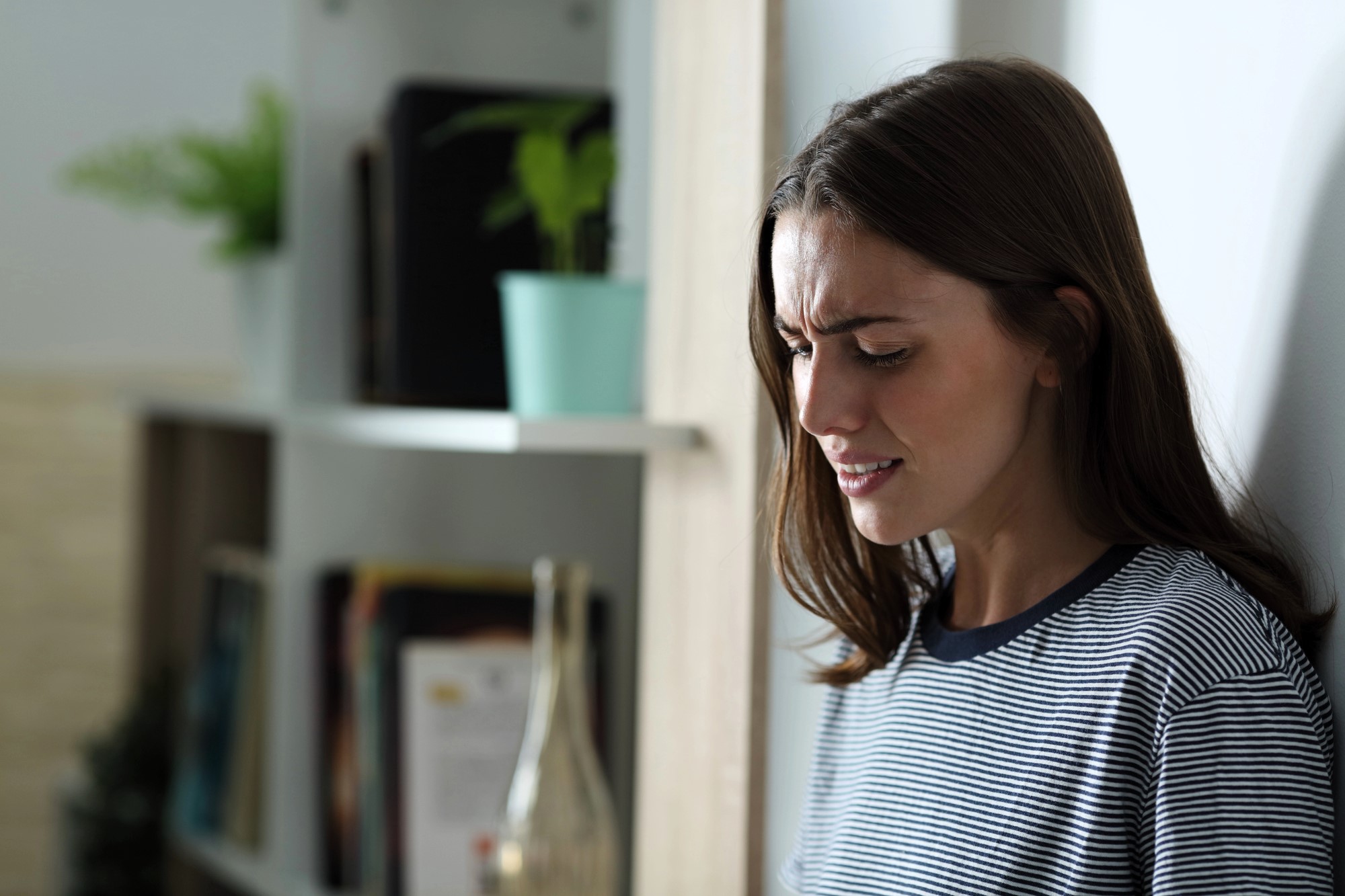 A woman in a striped shirt stands against a wall, looking concerned. The background features a bookshelf with plants and decorative items.