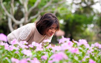 A woman with short hair is leaning over a field of blooming pink flowers, appearing to enjoy their scent. She is wearing a light-colored sweater. The background features green, blurred trees.