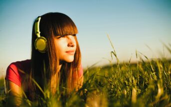 A young woman with long brown hair and bangs lies in tall grass, wearing yellow headphones. She gazes thoughtfully into the distance, bathed in warm sunlight. The sky is clear, creating a serene and peaceful atmosphere.