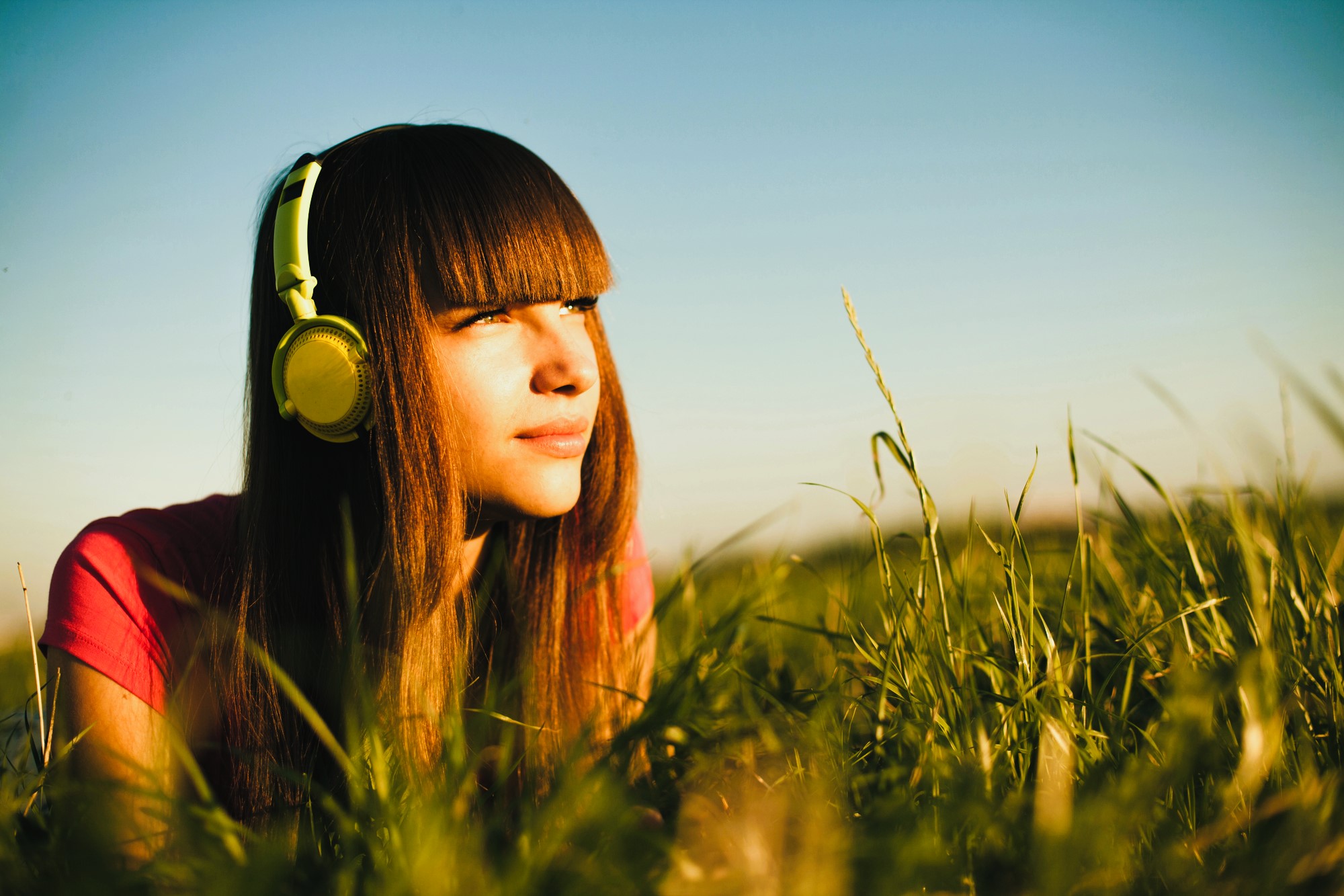 A young woman with long brown hair and bangs lies in tall grass, wearing yellow headphones. She gazes thoughtfully into the distance, bathed in warm sunlight. The sky is clear, creating a serene and peaceful atmosphere.