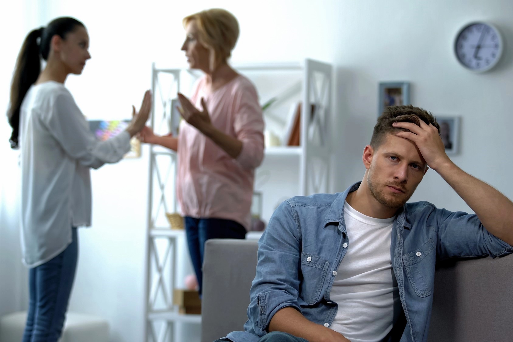 A tired man sits on a couch, resting his head on his hand, while two women in the background have an animated discussion. One woman gestures emphatically, and the other holds up a hand as if to stop the conversation. A wall clock shows 3:00.