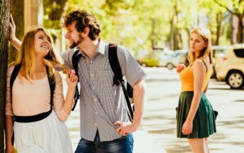 A couple stands outside, playful and smiling at each other. The man holds the woman's braid. Nearby, another woman in an orange top looks on with a neutral expression. The setting is a sunlit, tree-lined street with parked cars.