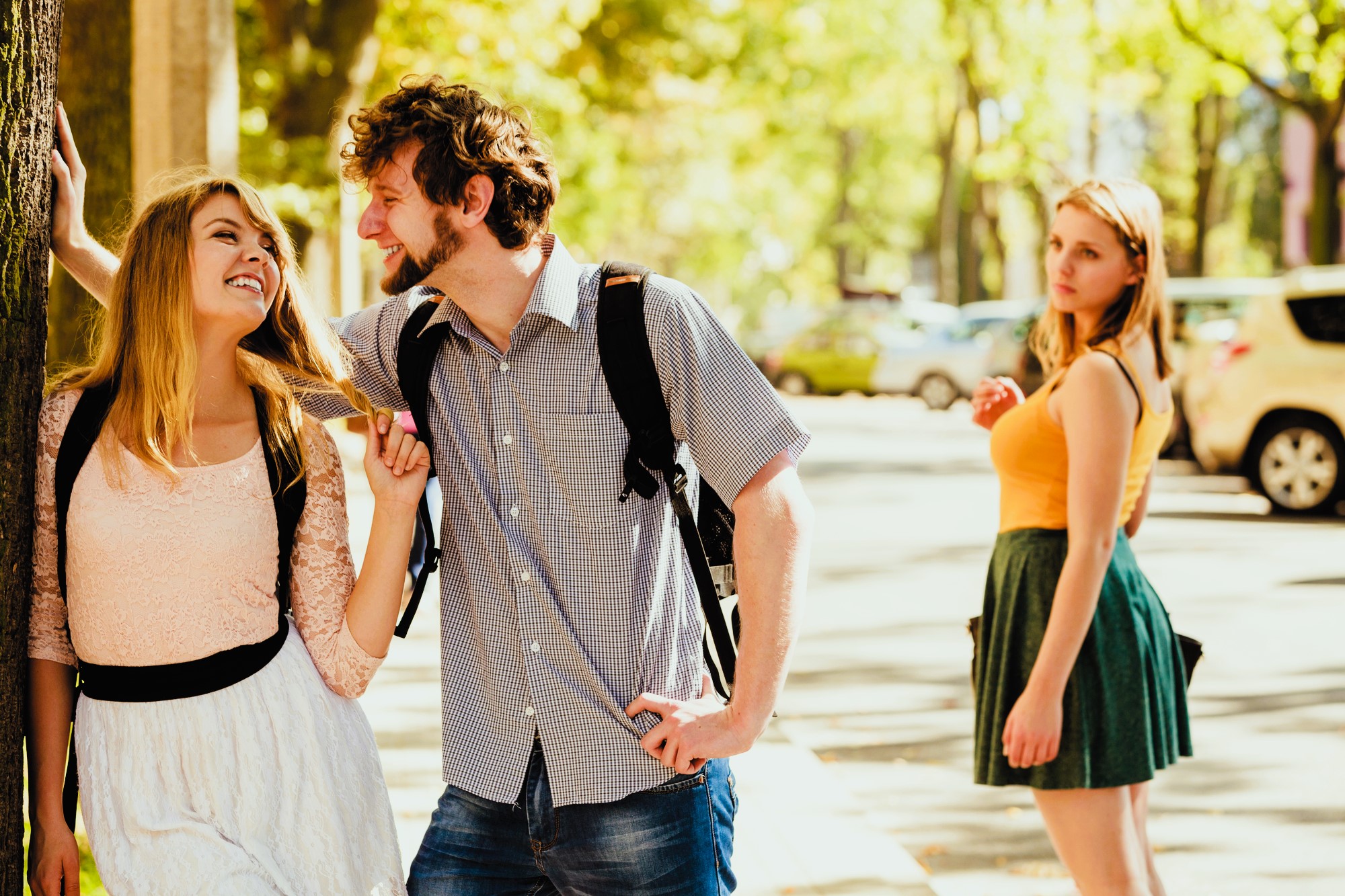 A couple stands outside, playful and smiling at each other. The man holds the woman's braid. Nearby, another woman in an orange top looks on with a neutral expression. The setting is a sunlit, tree-lined street with parked cars.