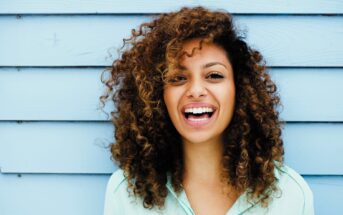 Smiling person with curly hair stands against a blue wooden wall, wearing a light blue shirt.