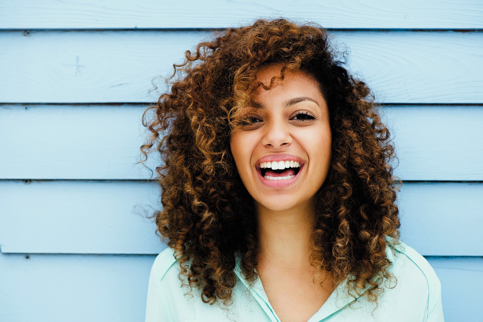 Smiling person with curly hair stands against a blue wooden wall, wearing a light blue shirt.