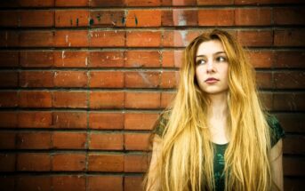 Woman with long blonde hair wearing a dark green top stands against a brick wall, looking to her right with a thoughtful expression. The wall has a few marks and scratches, adding texture to the background.