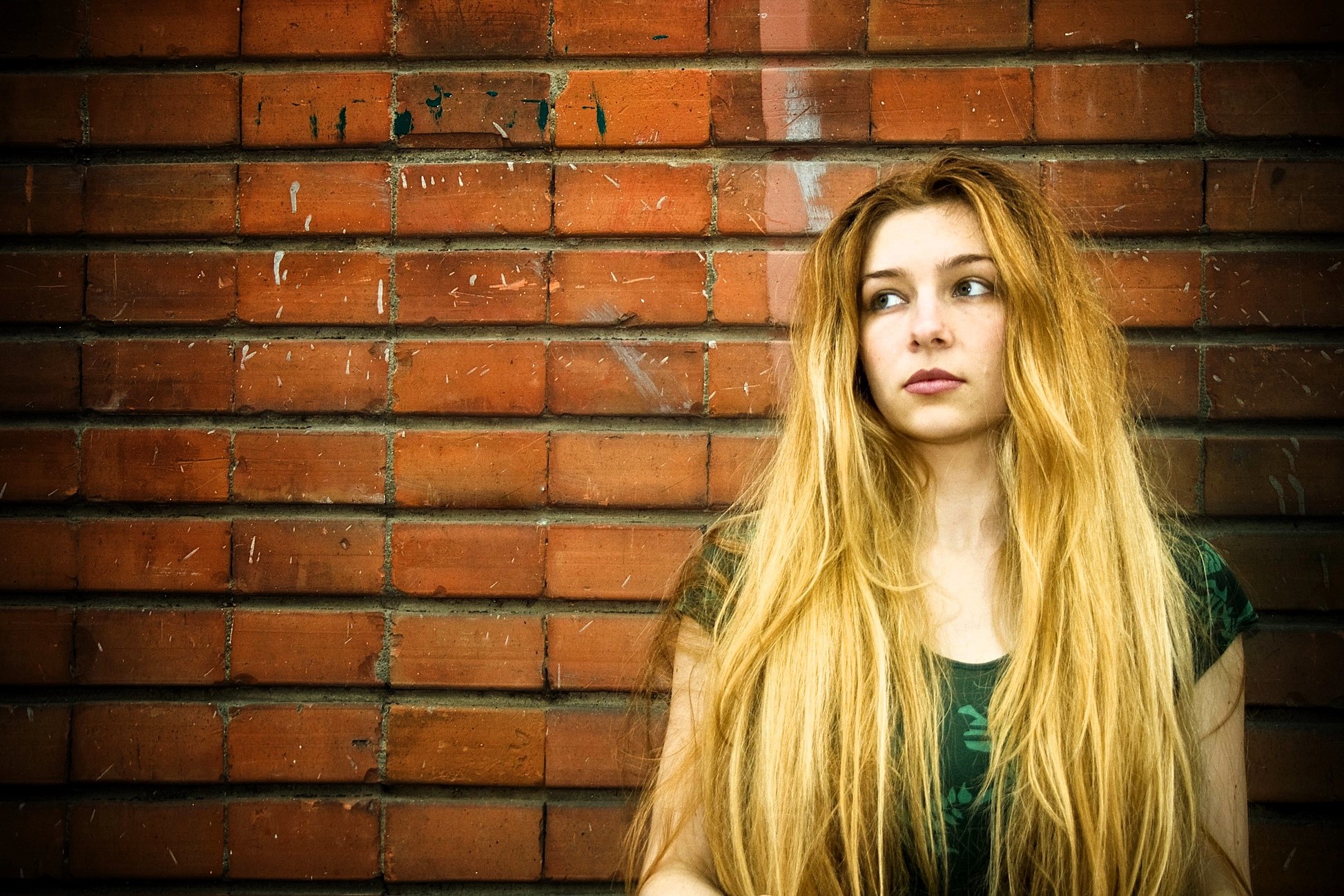 Woman with long blonde hair wearing a dark green top stands against a brick wall, looking to her right with a thoughtful expression. The wall has a few marks and scratches, adding texture to the background.