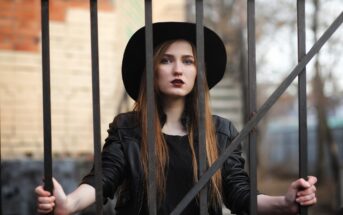 A woman with long brown hair stands behind a metal fence, holding the bars with both hands. She is wearing a wide-brimmed black hat and a black leather jacket. The background is blurred, showing a brick wall and trees.