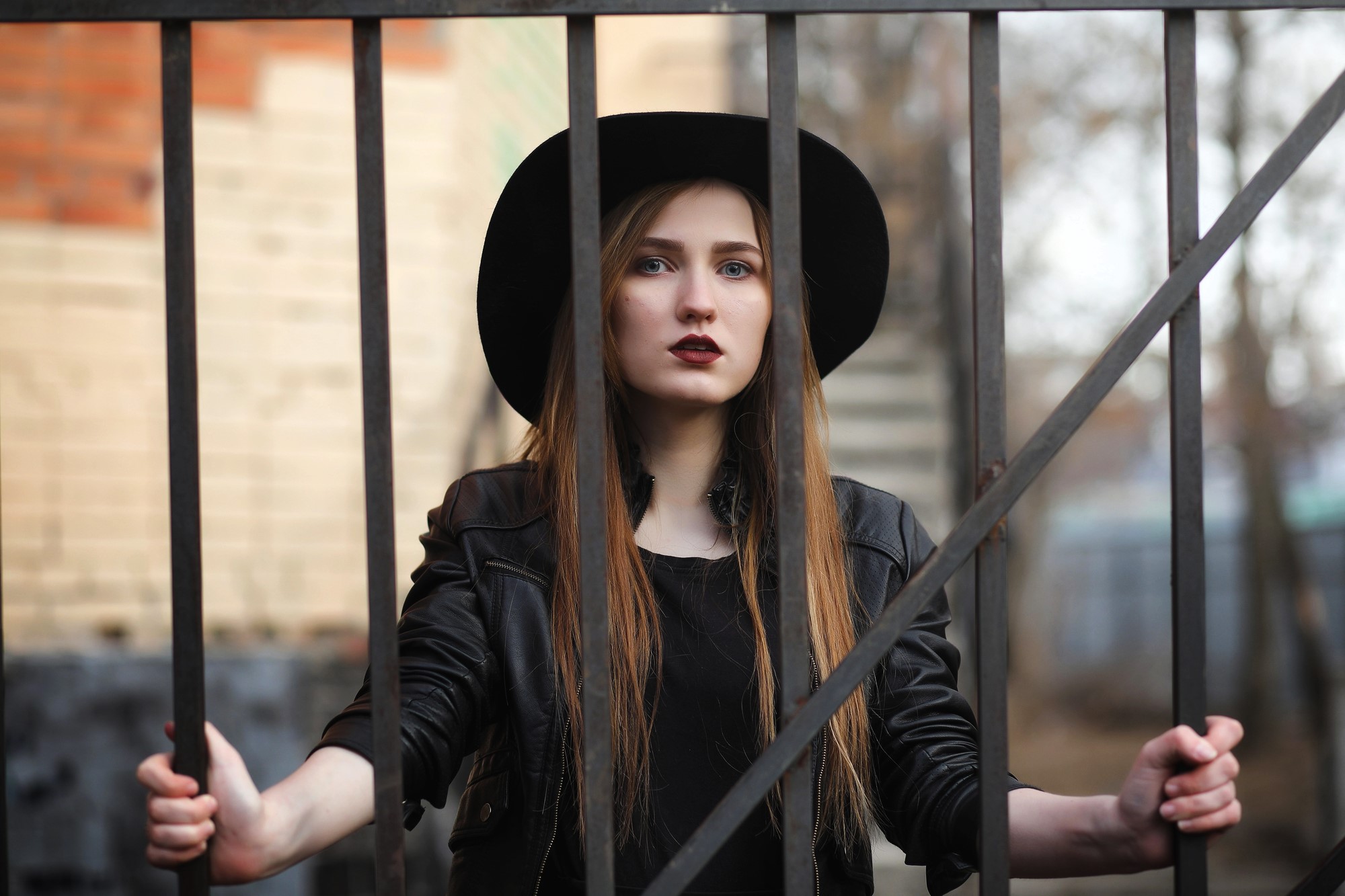 A woman with long brown hair stands behind a metal fence, holding the bars with both hands. She is wearing a wide-brimmed black hat and a black leather jacket. The background is blurred, showing a brick wall and trees.