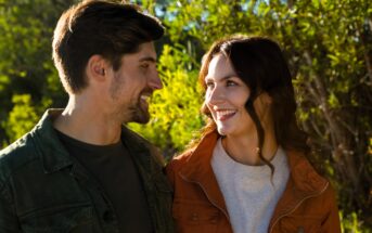 A man and woman, both smiling and wearing casual jackets, stand together outdoors. The sunlight illuminates their faces, and there is greenery in the background, suggesting a warm, sunny day.