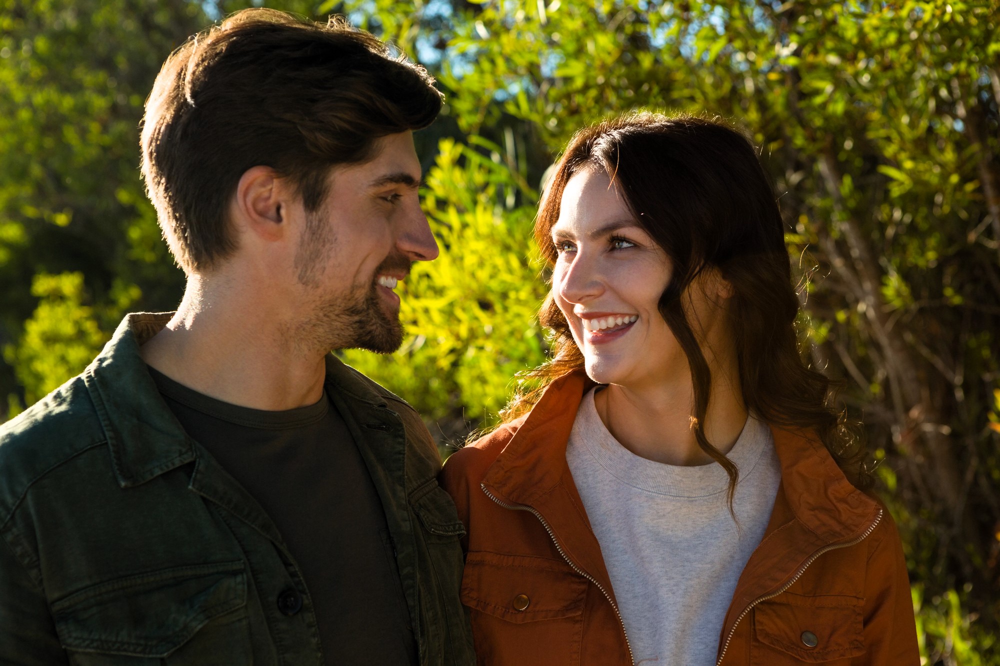 A man and woman, both smiling and wearing casual jackets, stand together outdoors. The sunlight illuminates their faces, and there is greenery in the background, suggesting a warm, sunny day.