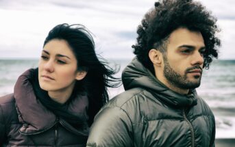 A woman and a man stand closely together outdoors, both wearing puffer jackets. The ocean is visible in the background. The woman's hair flows in the wind, while the man looks into the distance with a serious expression. The sky is cloudy.