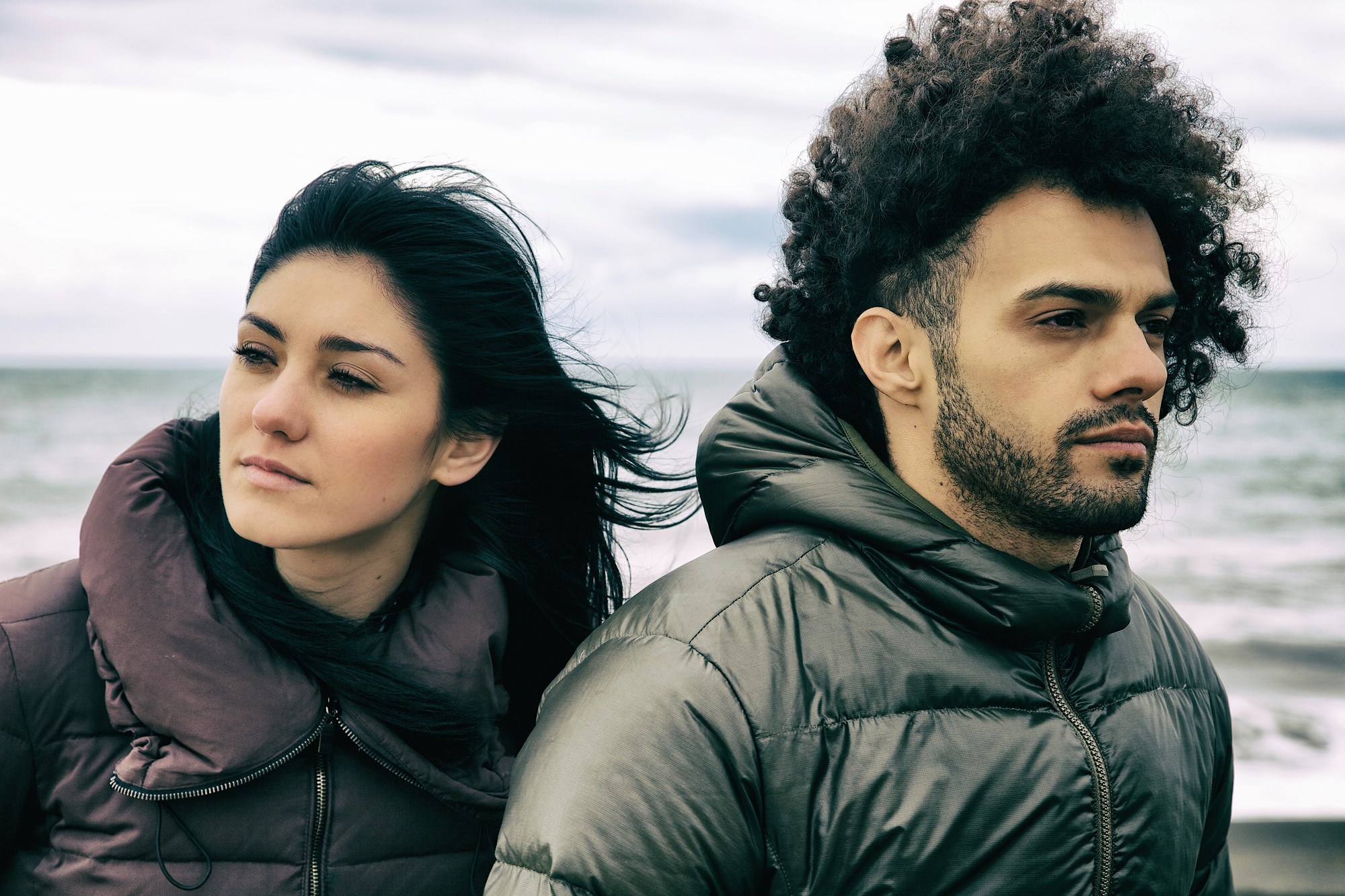 A woman and a man stand closely together outdoors, both wearing puffer jackets. The ocean is visible in the background. The woman's hair flows in the wind, while the man looks into the distance with a serious expression. The sky is cloudy.