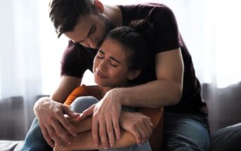 A man with a beard sits closely, embracing a woman who appears comforted yet emotional with closed eyes. They are seated on a couch with sunlight filtering through a window in the background.