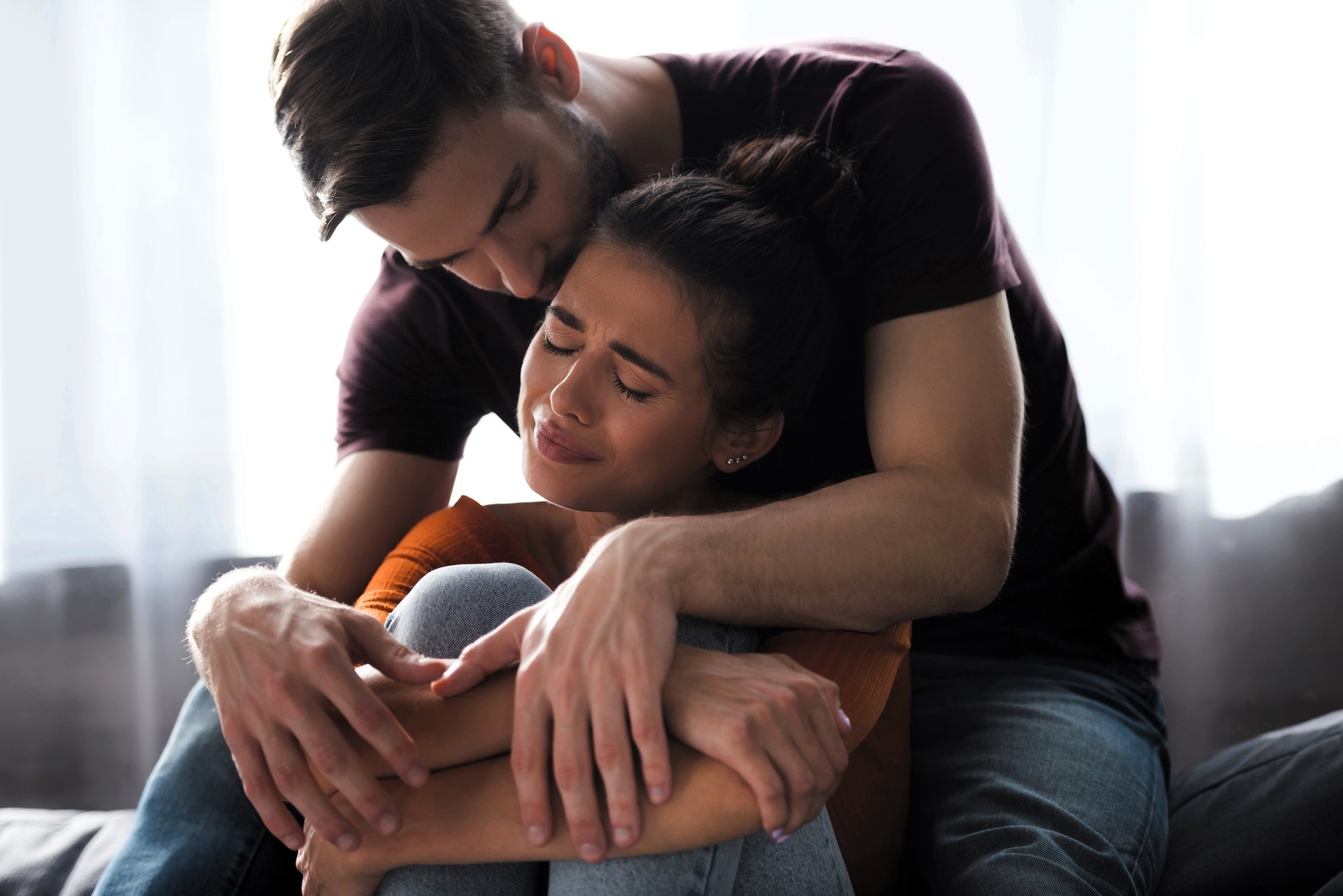 A man with a beard sits closely, embracing a woman who appears comforted yet emotional with closed eyes. They are seated on a couch with sunlight filtering through a window in the background.